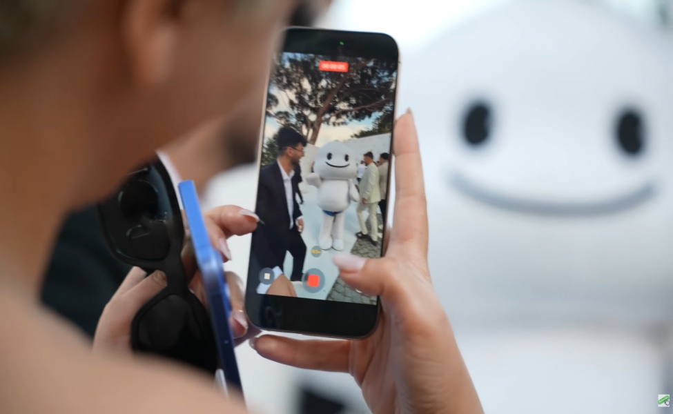 Person holding smartphone recording video of white robot character outdoors